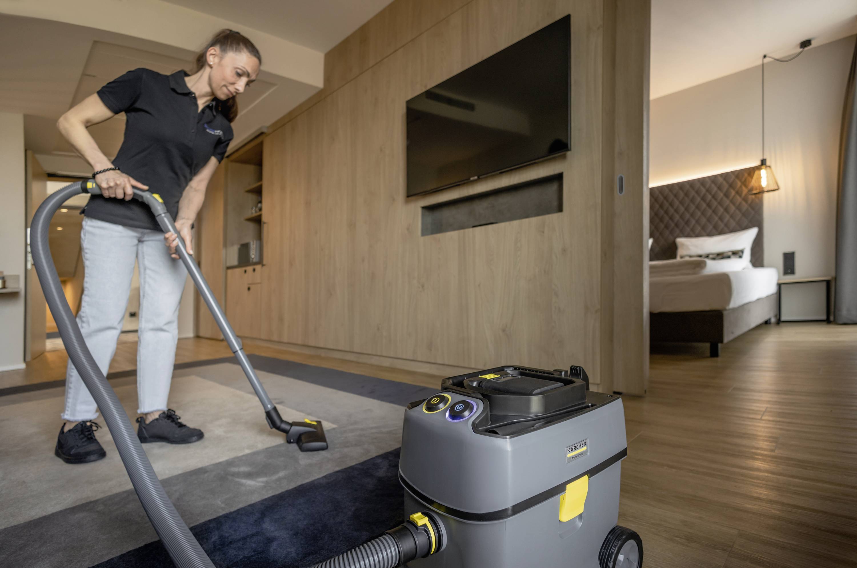 Eine Frau staubsaugt den Teppich in einem modernen Zimmer mit Holzdekor und einem Bett im Hintergrund. Sie trägt ein dunkles Poloshirt und Jeans.