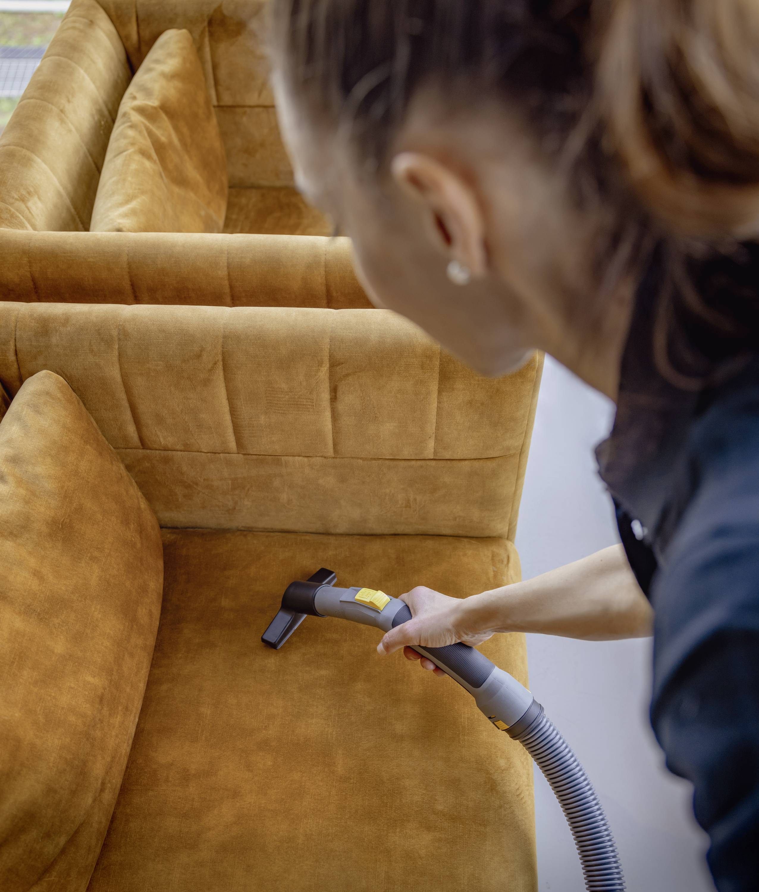 Eine Person reinigt mit einem Staubsauger das Polster eines braunen Sofas. Die Nahaufnahme zeigt den Staubsauger in Aktion.