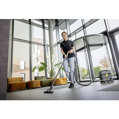 Eine Person saugt den Boden in einem modernen, lichtdurchfluteten Gebäude. Im Hintergrund sind Pflanzen und Sitzmöbel zu sehen.