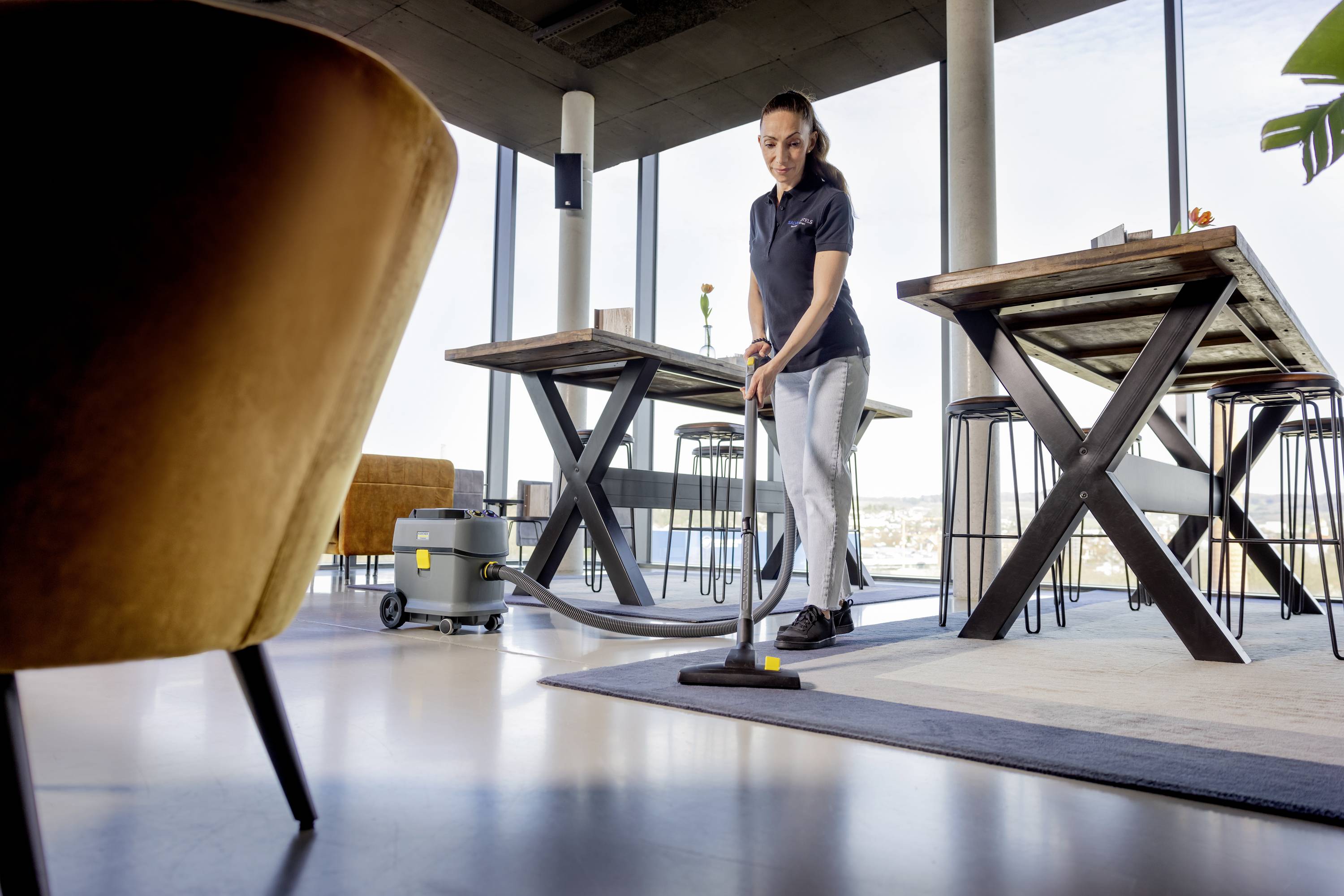 Eine Person reinigt den Boden eines modernen, hellen Büros mit einem Staubsauger. Im Hintergrund sind hohe Tische und Stühle sichtbar.