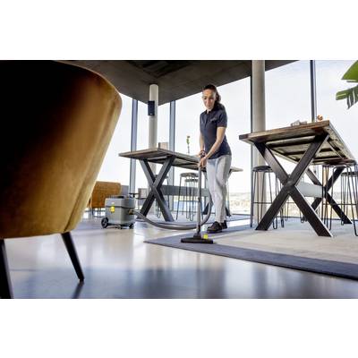 Eine Person reinigt den Boden eines modernen, hellen Büros mit einem Staubsauger. Im Hintergrund sind hohe Tische und Stühle sichtbar.
