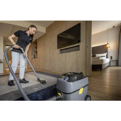 Eine Reinigungskraft staubsaugt den Teppich in einem modernen Hotelzimmer. Im Hintergrund sind ein Bett und Holzmöbel sichtbar.