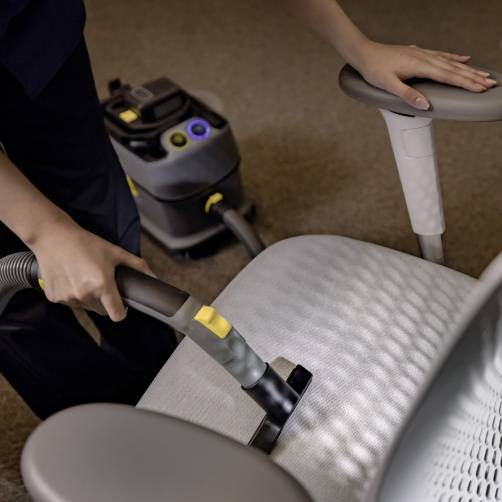 Eine Person reinigt den Sitz eines Stuhls mit einem Staubsauger. Im Hintergrund steht ein weiterer Staubsauger mit Zubehör.
