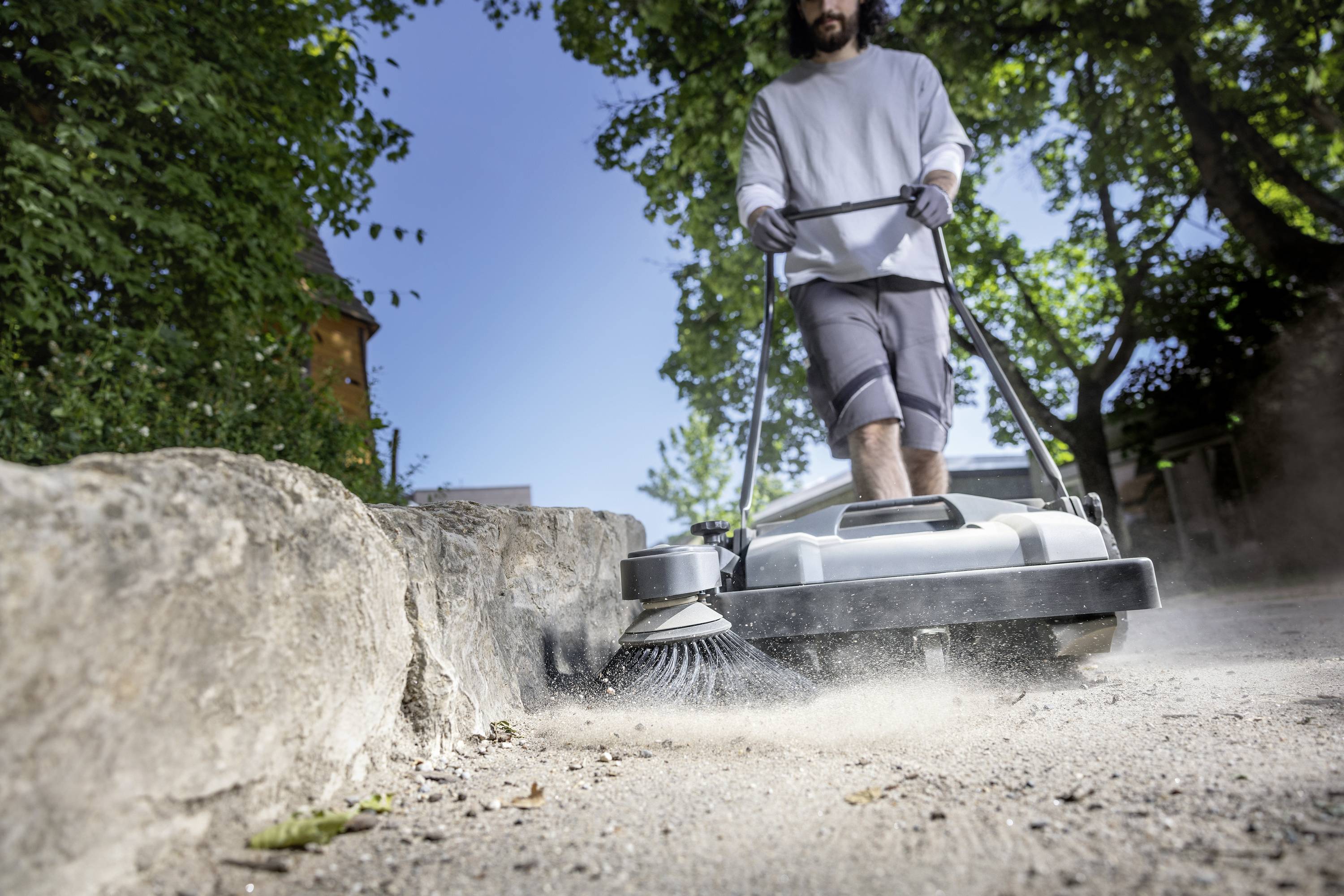 Eine Person fegt mit einer Kehrmaschine Staub und Schmutz auf einem Gehweg. Bäume und Gebäude sind im Hintergrund zu sehen.