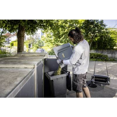 Eine Person entsorgt Pappe in einen großen Müllcontainer im Freien. Im Hintergrund sind Bäume und ein Gehweg zu sehen.