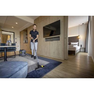 Eine Person saugt einen Teppich in einem modernen Hotelzimmer. Im Hintergrund ist ein Bett sichtbar.
