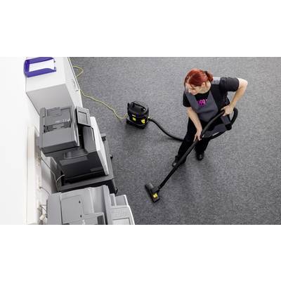 Eine Person mit einem Staubsauger reinigt einen Teppichboden in einem Büro mit mehreren Kopiergeräten an einer Wand.