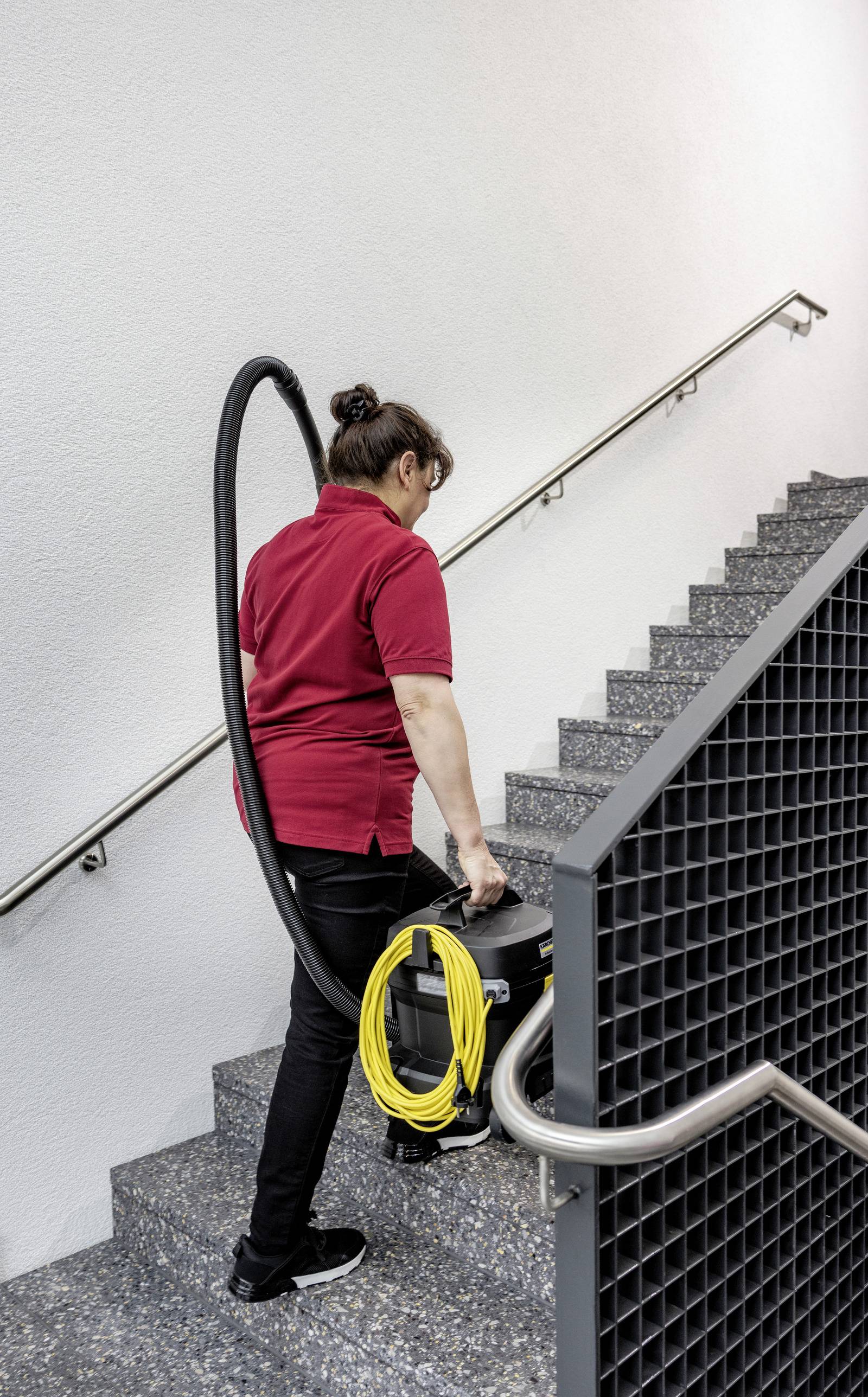 Eine Person in einem roten Hemd trägt einen Staubsauger mit einem gelben Kabel eine graue Treppe hinauf und deutet damit auf Reinigungsaktivitäten hin.