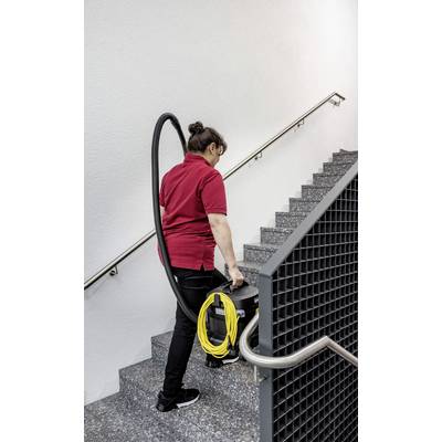 Eine Person in einem roten Hemd trägt einen Staubsauger mit einem gelben Kabel eine graue Treppe hinauf und deutet damit auf Reinigungsaktivitäten hin.