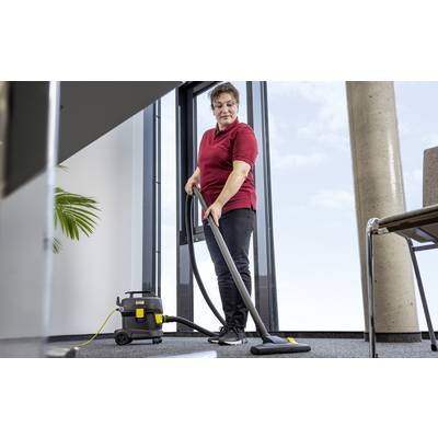 Eine Person in einem roten Hemd saugt einen Teppichboden in einem modernen Büroraum, wobei ein Fenster und eine Pflanze im Hintergrund sichtbar sind.