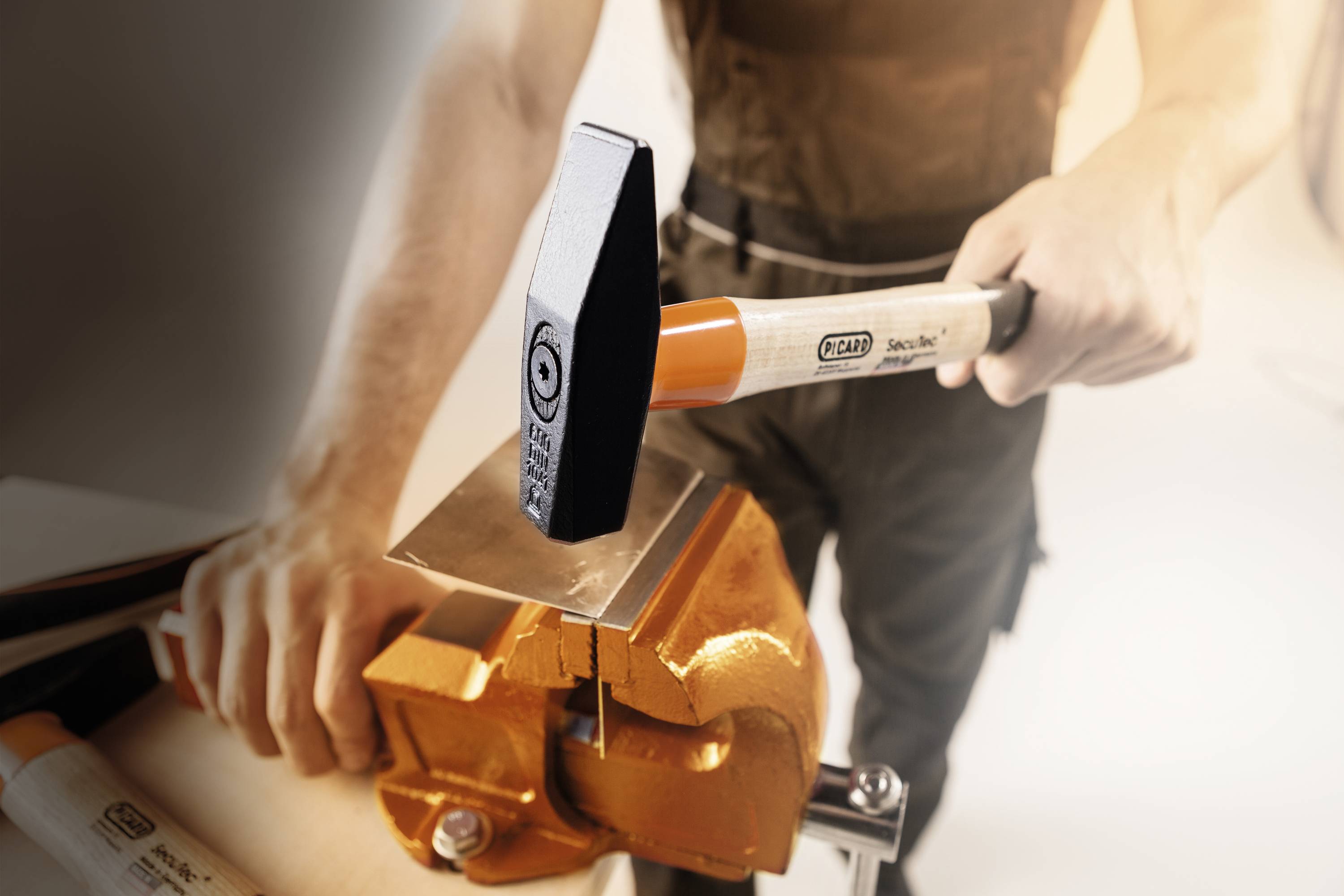 Eine Person schlägt mit einem Hammer auf ein Metallstück, das in einem Schraubstock eingespannt ist. Sie trägt Arbeitskleidung.