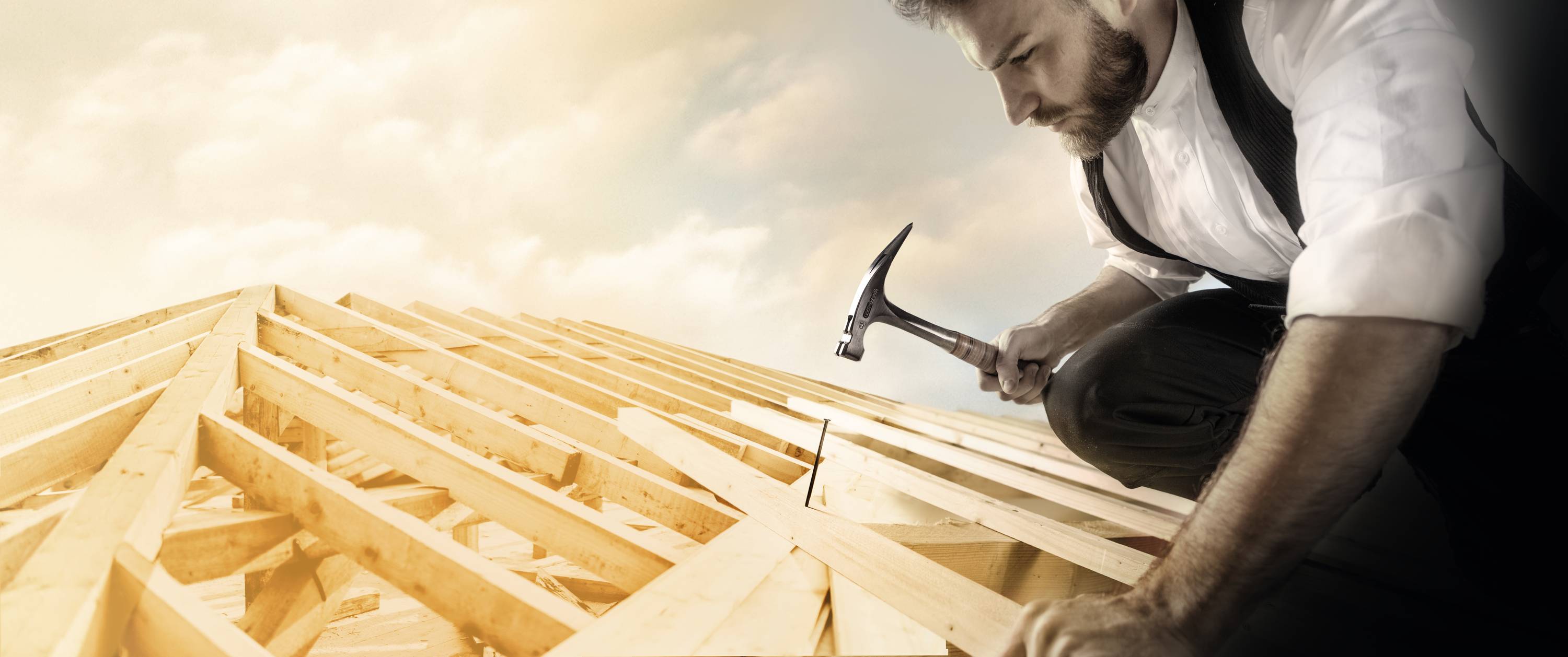 Ein Mann arbeitet konzentriert auf einem Dachstuhl aus Holz, hält einen Hammer in der Hand unter einem bewölkten Himmel.