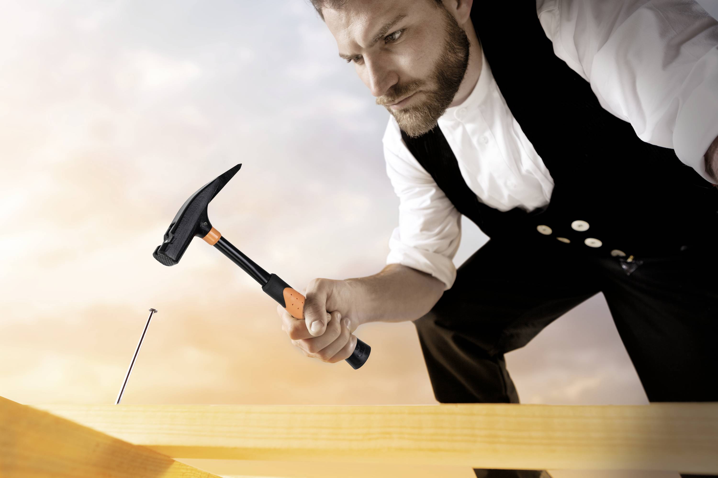 Ein Mann in traditioneller Handwerkskleidung benutzt einen Hammer, um einen Nagel in ein Holzbrett zu schlagen, bei Sonnenuntergang.