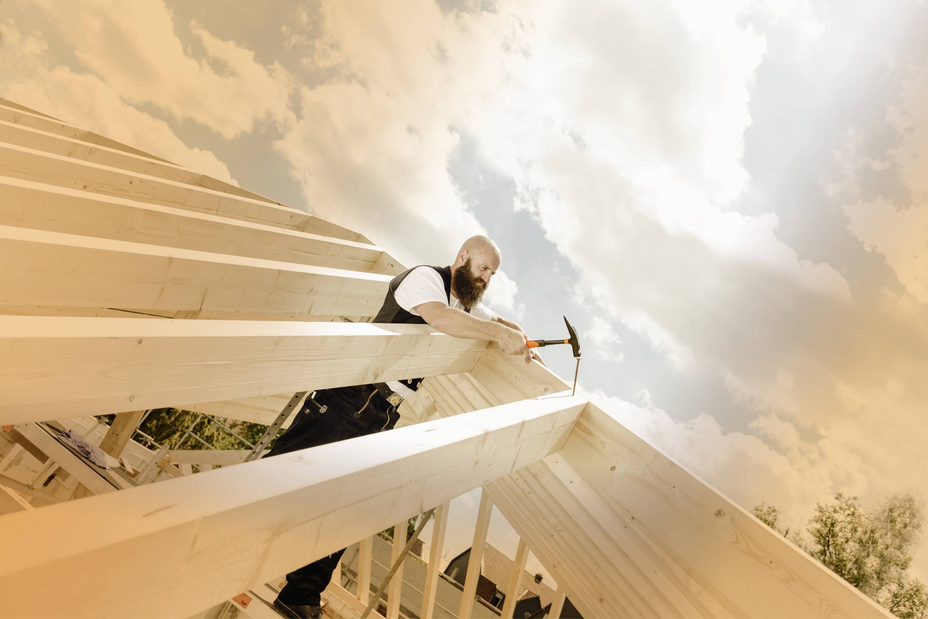 Ein Mann arbeitet auf einem Holzgerüst unter bewölktem Himmel. Er hält einen Hammer und befestigt Teile des Holzdachs.