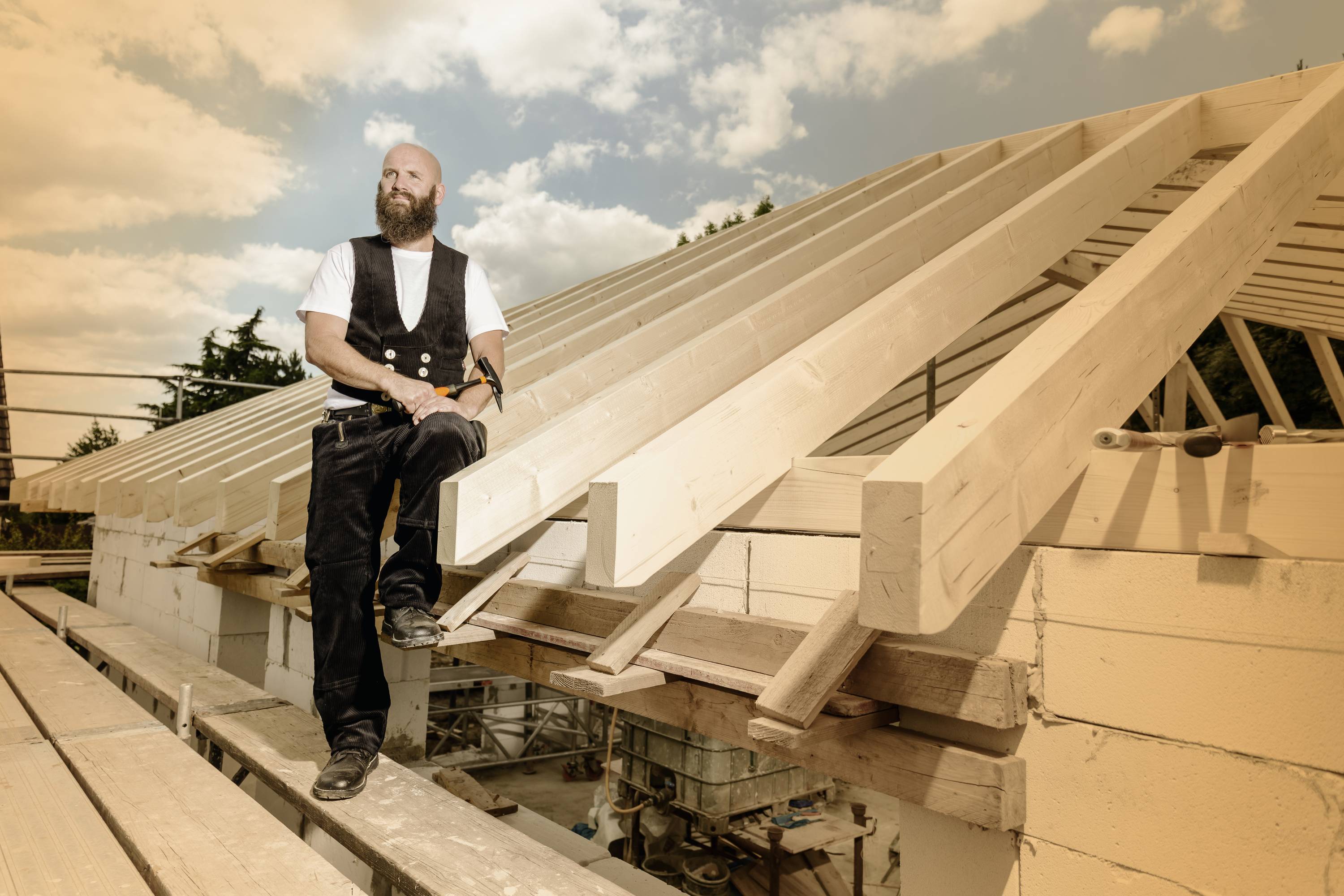 Ein Mann im Zimmermannsoutfit steht auf einem Hausdach in Bau, umgeben von Holzbalken und einem bewölkten Himmel.