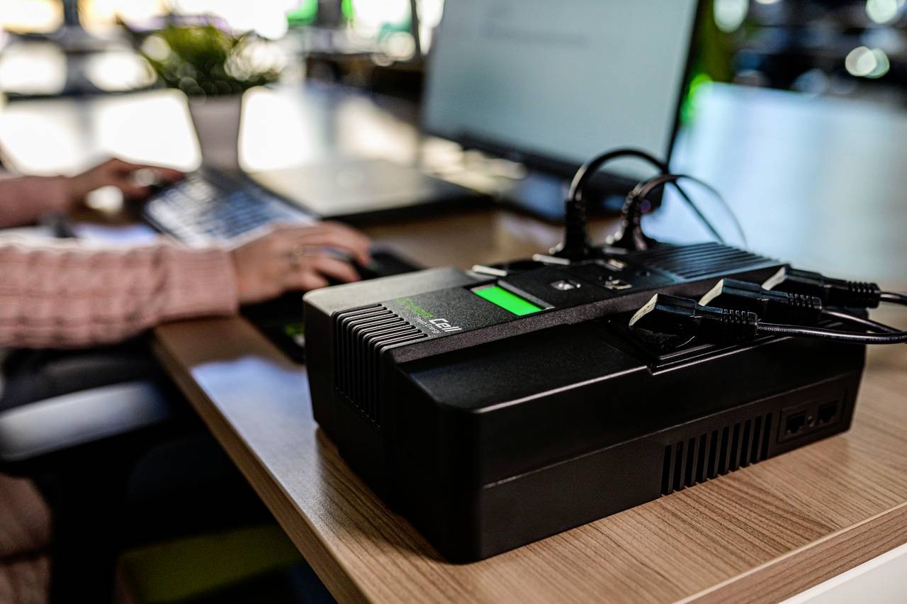 Eine Person arbeitet an einem Laptop, während im Vordergrund ein Gerät mit mehreren Kabeln auf einem Tisch steht.