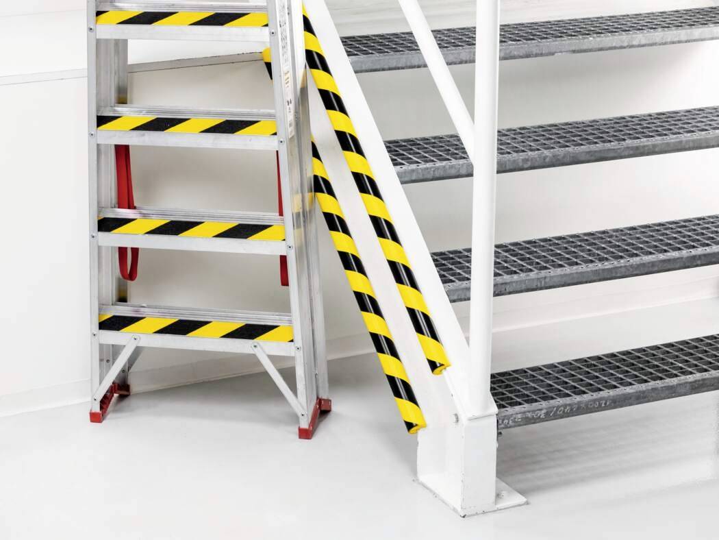 'Metalltreppe mit schwarz-gelben Sicherheitsstreifen an Geländer und Stufen, roter Handlauf, weiße Wände, grauer Boden.'