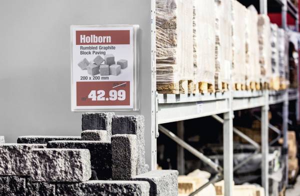 'Holborn Rumbled Graphite Block Paving' Schild zeigt Maße 200 x 200 mm und Preis 42,99 pro Quadratmeter neben Stapeln von Pflastersteinen.