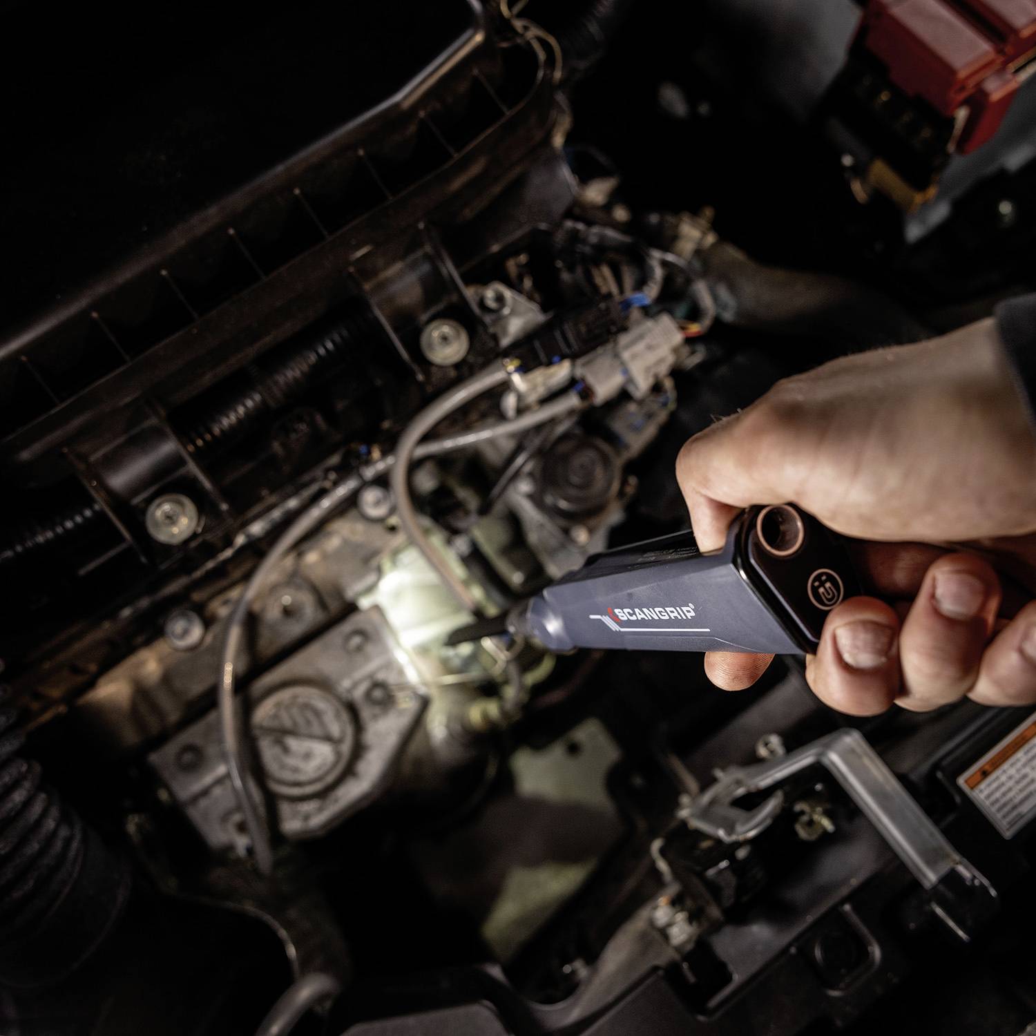 Eine Person prüft den Motor eines Autos mit einem Handgerät, wahrscheinlich zur Diagnose. Kabel und Motorteile sind sichtbar.