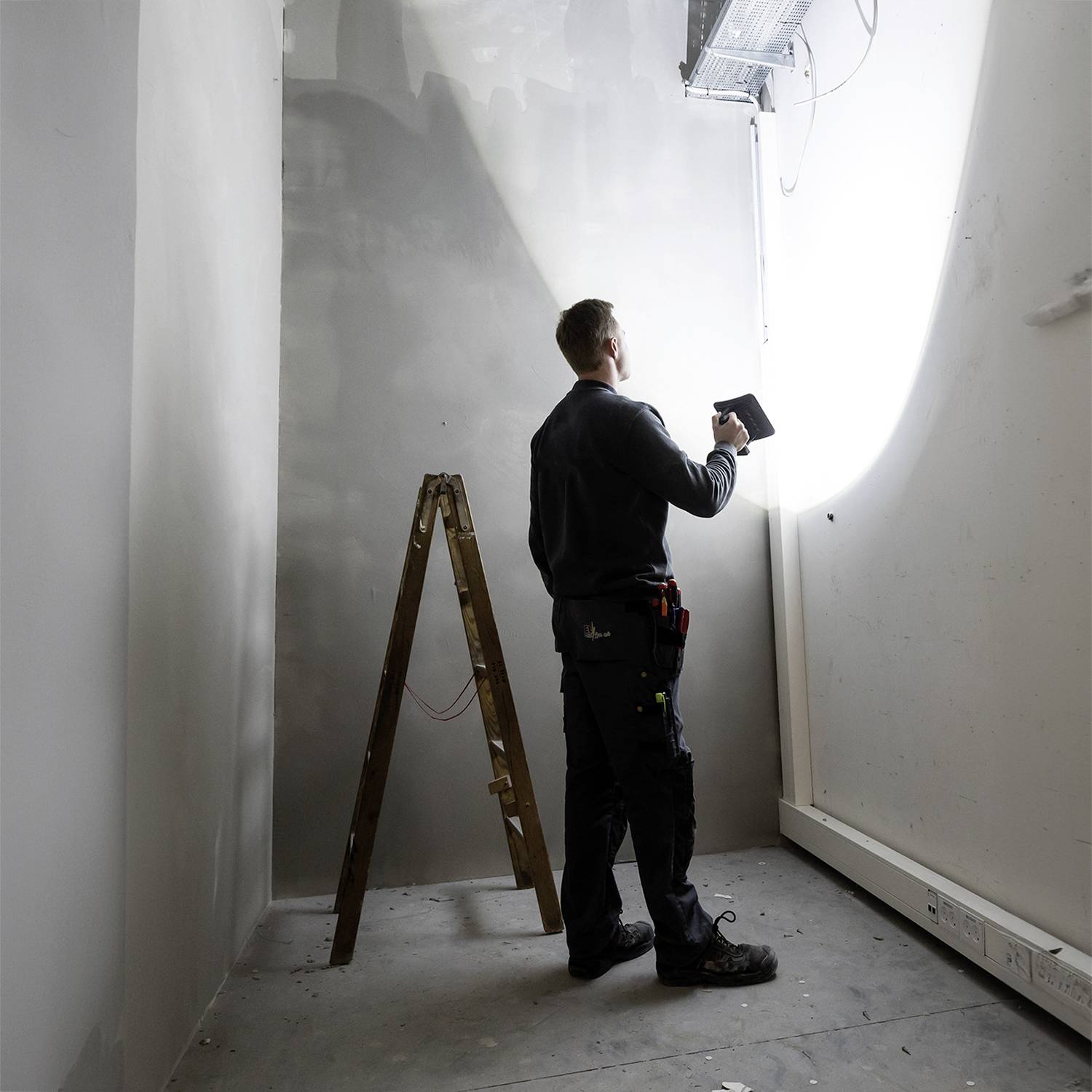 Ein Handwerker steht in einem schmalen Raum und beleuchtet die Wand mit einer Lampe. Eine Holzleiter steht in der Nähe.