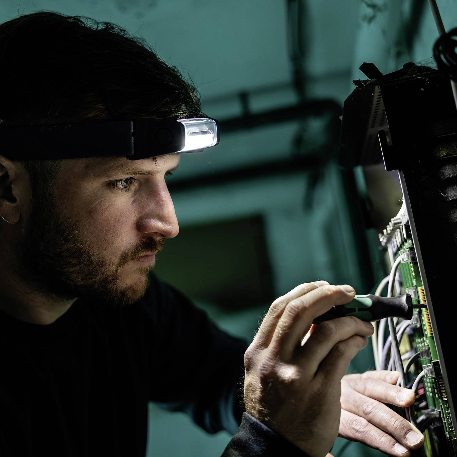 Ein Elektriker trägt ein Stirnlicht und benutzt einen Schraubendreher, um an einer geöffneten Schalttafel in einem schlecht beleuchteten Raum zu arbeiten.