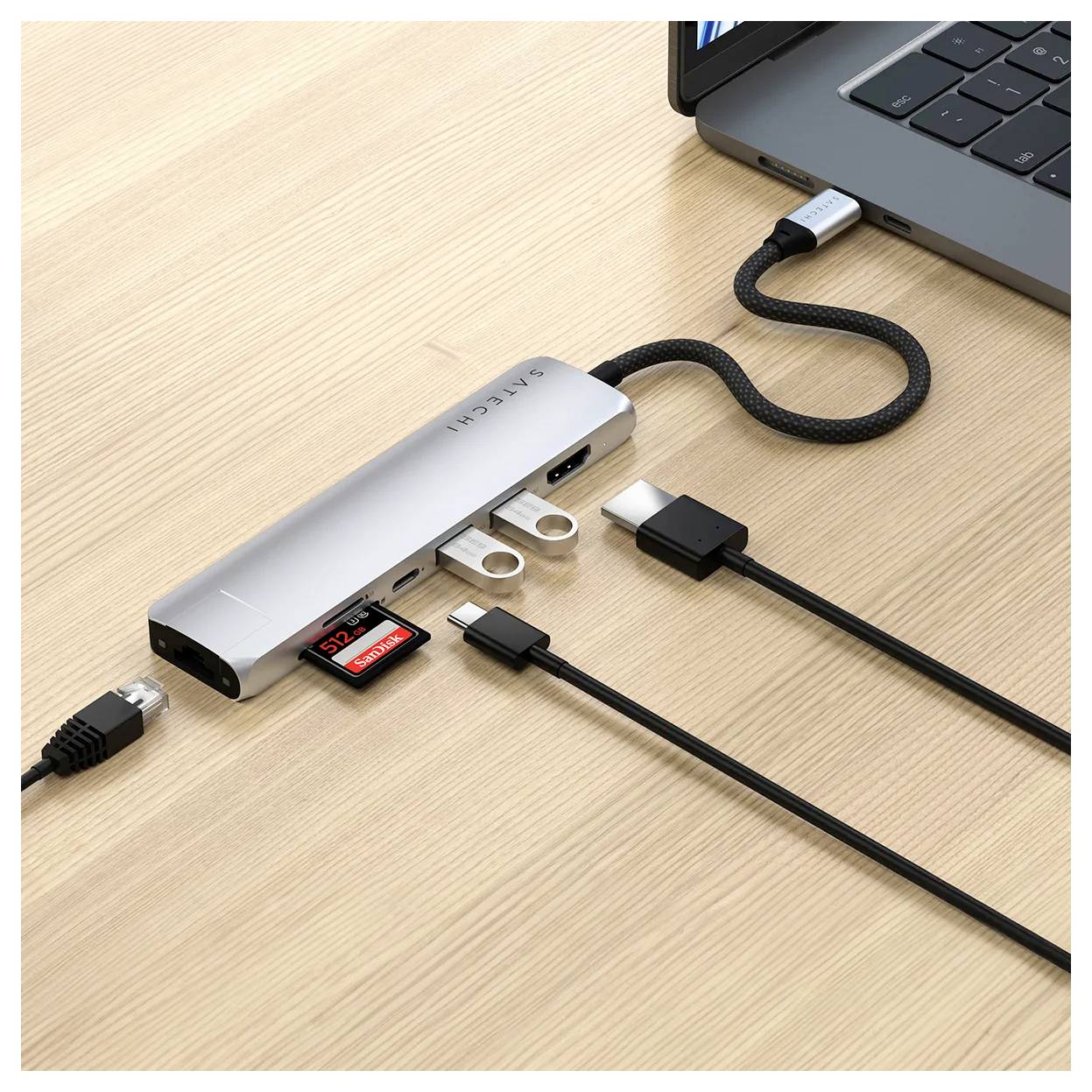Ein Laptop ist mit einem USB-C-Hub verbunden, der mehrere Anschlüsse bietet, darunter HDMI, USB und Kartenleser. Auf einem Holztisch.
