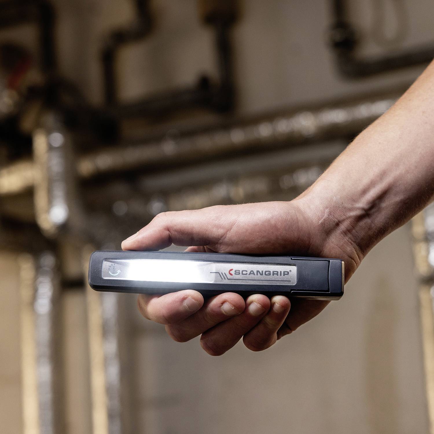 Eine Hand hält eine längliche Taschenlampe vor einem unscharfen Hintergrund mit Rohren.