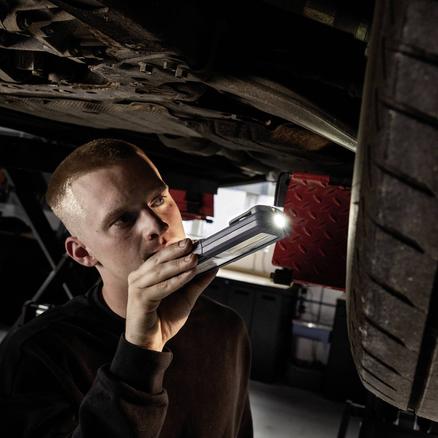Ein Mechaniker inspiziert das Fahrwerk eines Fahrzeugs mit einer Lampe in der Werkstatt. Die Szene zeigt konzentrierte Arbeit.