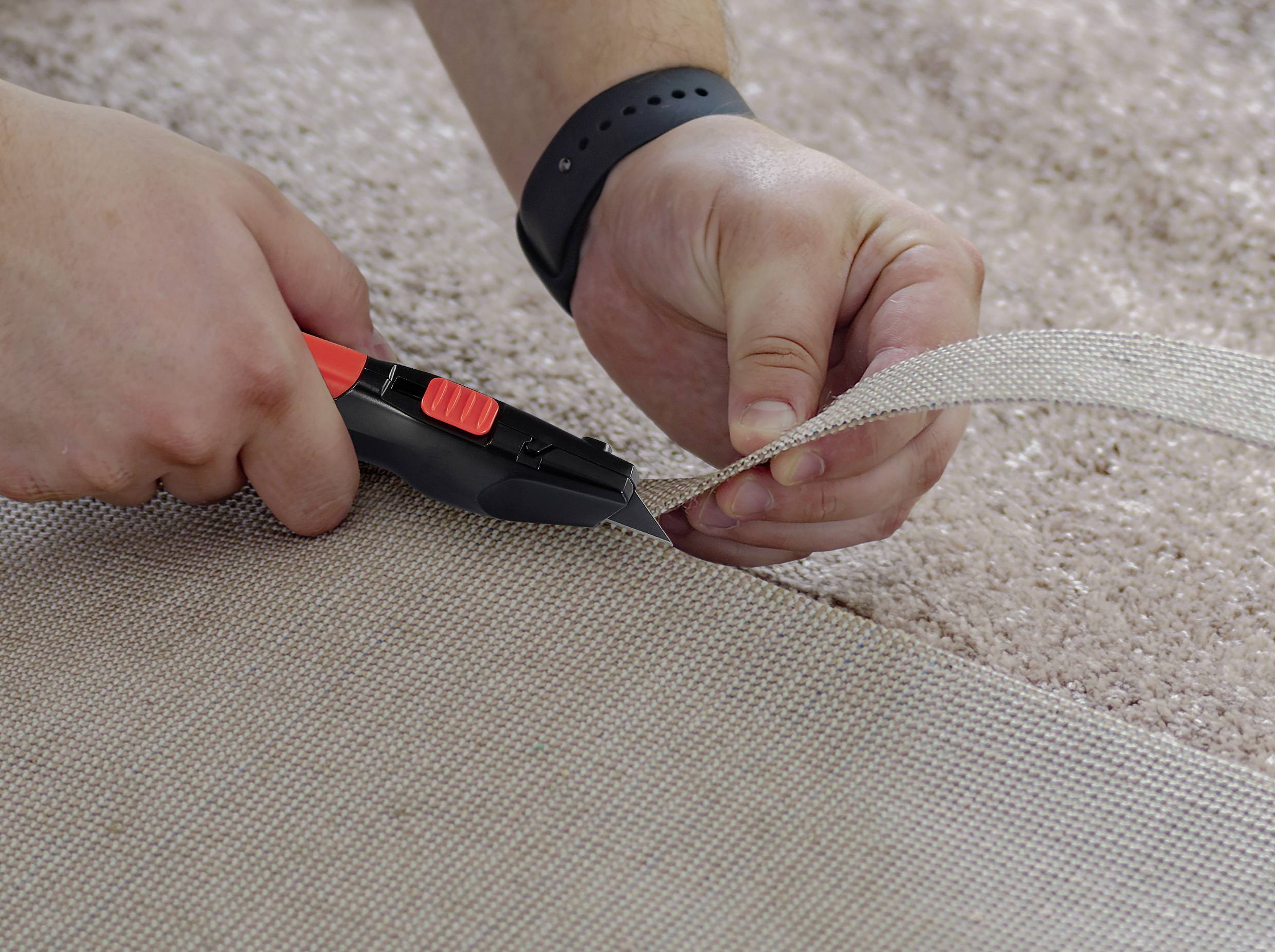 'Zwei Hände schneiden Teppich mit einem Cuttermesser auf einem beige Teppichboden. Man trägt ein schwarzes Armband.'