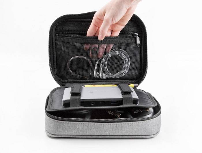 Eine geöffnete Reisetasche aus Stoff mit Fächern, die elektronische Geräte und Kabel ordentlich aufbewahrt. Eine Hand greift nach einem Kabel.