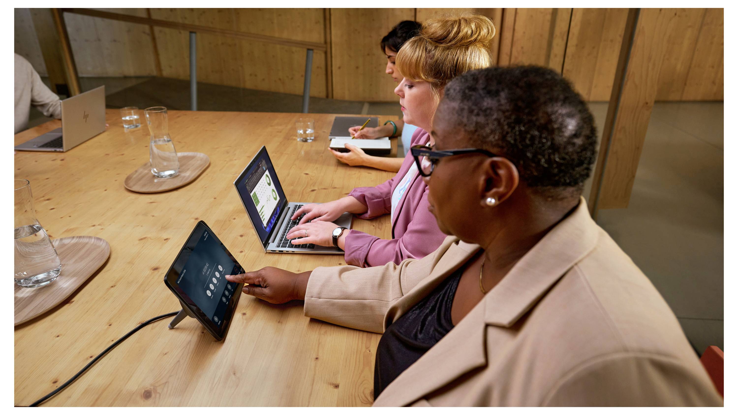 Drei Personen sitzen an einem Konferenztisch mit Laptops. Eine Person bedient ein Touchpanel. Es ist eine Büroszene.