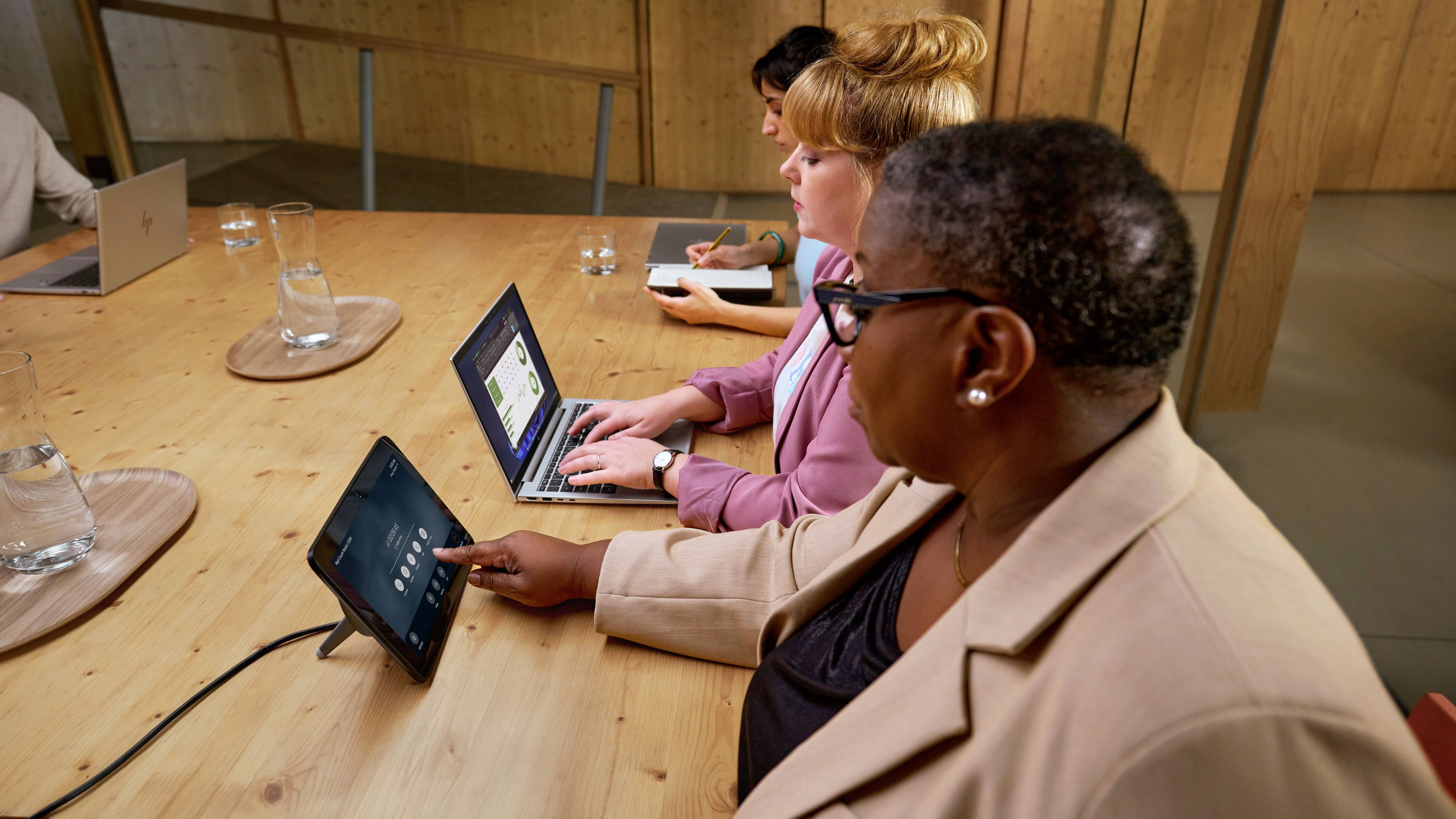 Drei Personen sitzen an einem Konferenztisch mit Laptops. Eine Person bedient ein Tablet, während die anderen Notizen machen.