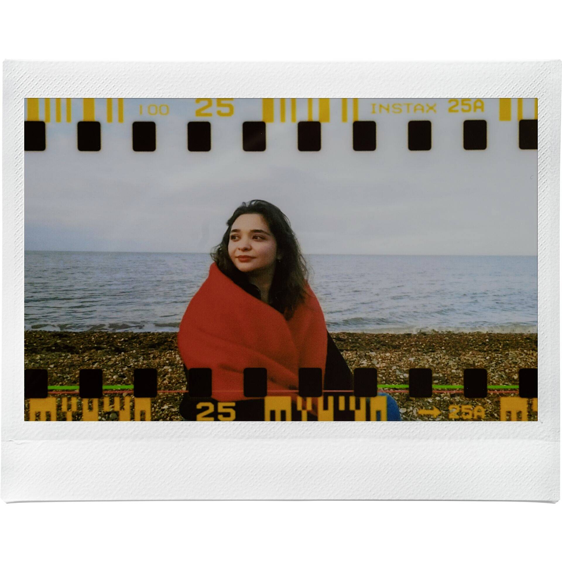 Eine Frau sitzt eingehüllt in einer roten Decke am Kiesstrand. Im Hintergrund ist das Meer zu sehen. Ein analoger Filmrahmen umgibt das Bild.