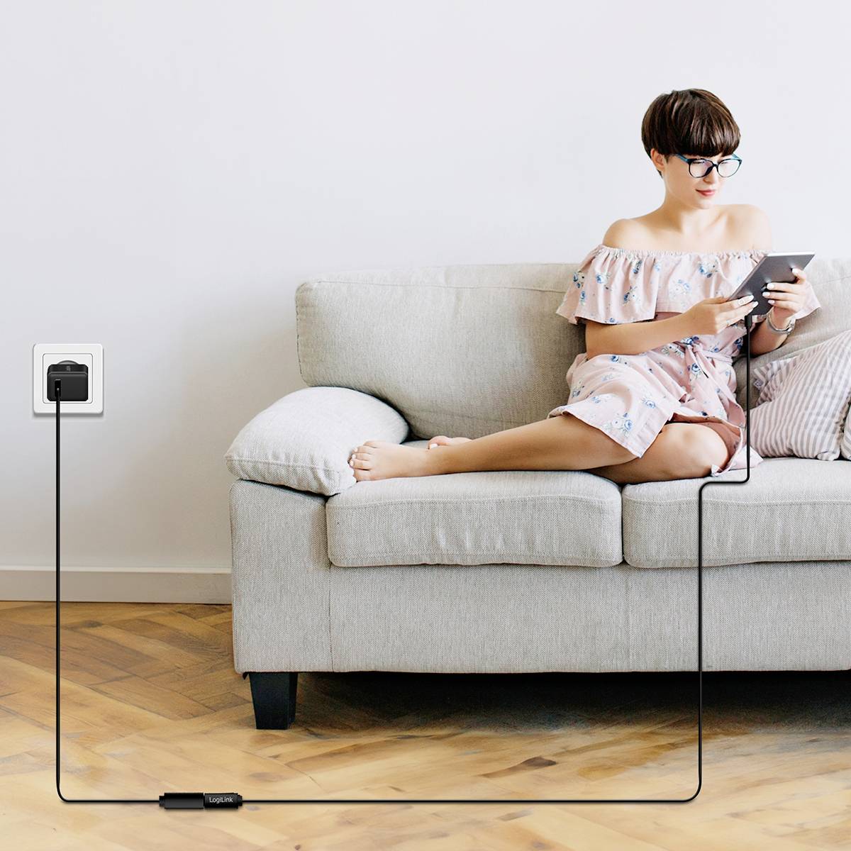 Frau sitzt auf einem Sofa und benutzt ein Smartphone, während das Gerät über ein langes Kabel mit einer Steckdose verbunden ist.