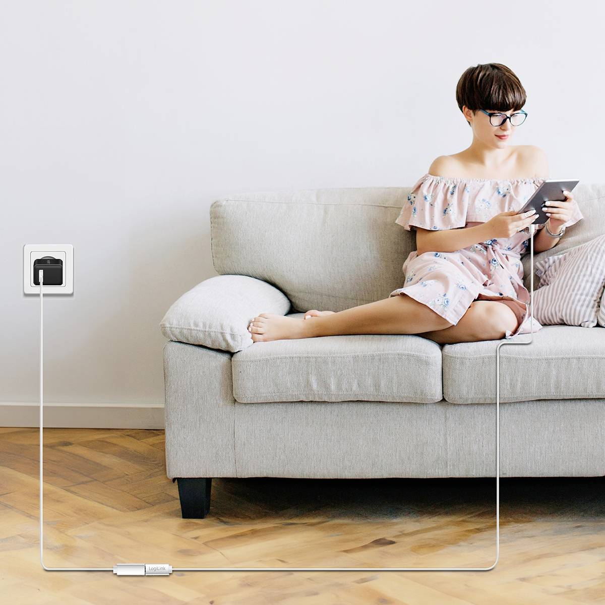 Eine Person sitzt auf einem hellen Sofa und liest aufmerksam ein Buch. Ein Ladekabel ist mit einer Steckdose an der Wand verbunden.