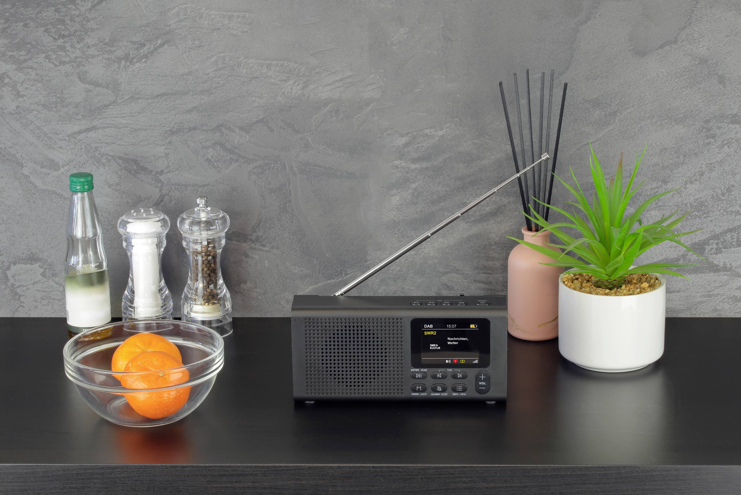 Ein schwarzes Radio steht auf einem Tisch neben einer Glasschale mit einer Orange, Salz- und Pfefferstreuer und einer Pflanze.