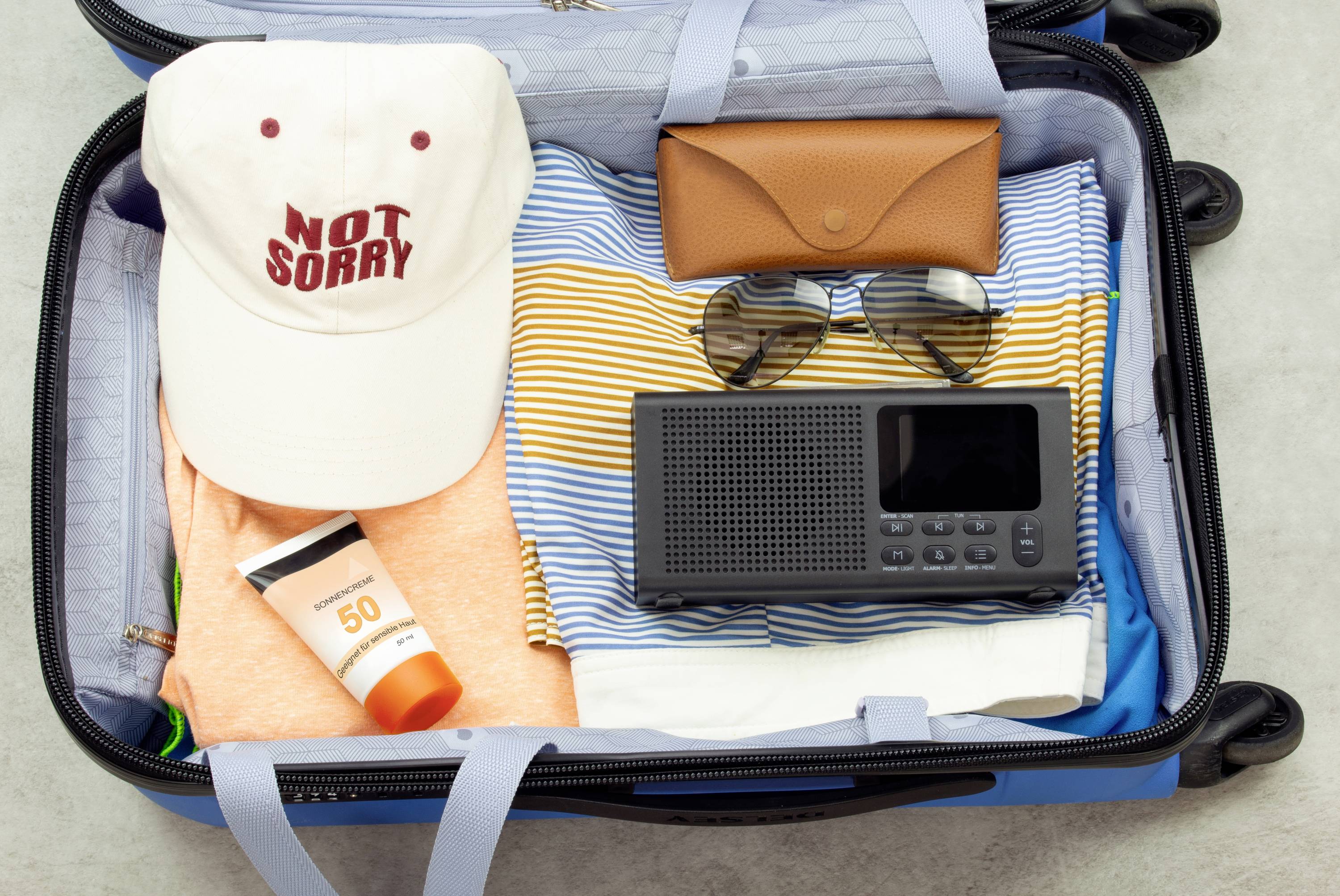 Ein geöffneter Koffer mit einem Hut, auf dem 'NOT SORRY' steht, Sonnenbrille, Sonnencreme, ein Radio und Kleidung sind zu sehen.