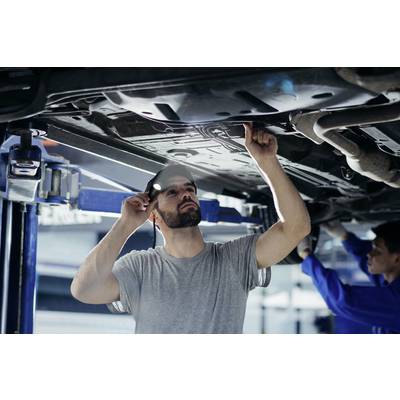 Ein Mechaniker inspiziert den Unterboden eines angehobenen Autos in einer Werkstatt, hält eine Taschenlampe und zeigt auf ein Bauteil.