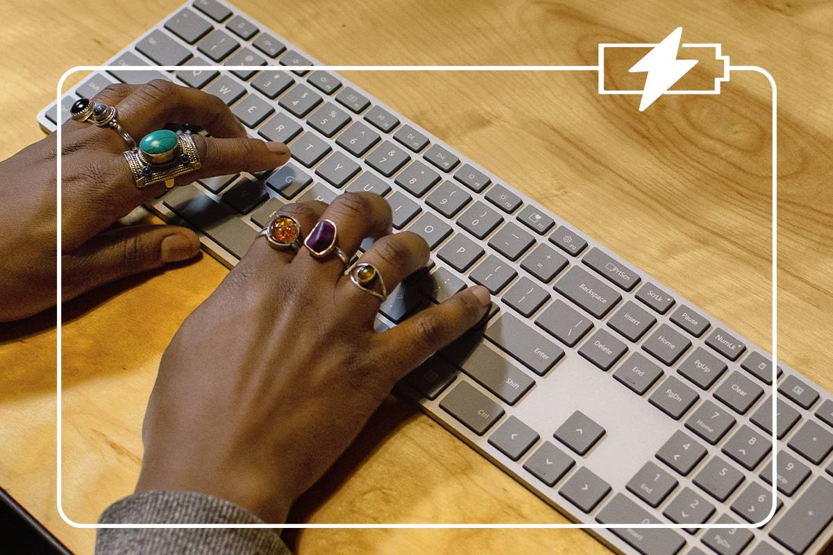 Hände tippen auf einer Computertastatur. Die Person trägt mehrere Ringe. In der Ecke oben rechts ist ein Akku-Symbol.