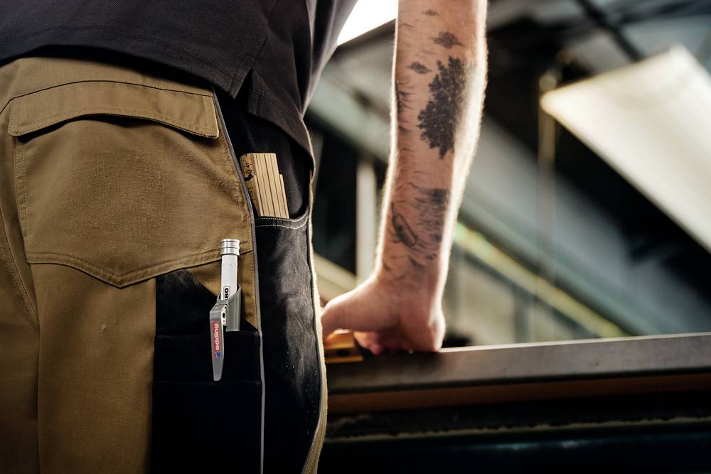 Ein Arbeiter mit tätowiertem Arm steht an einer Werkbank. Ein Stift und ein Block ragen aus der Hosentasche heraus.