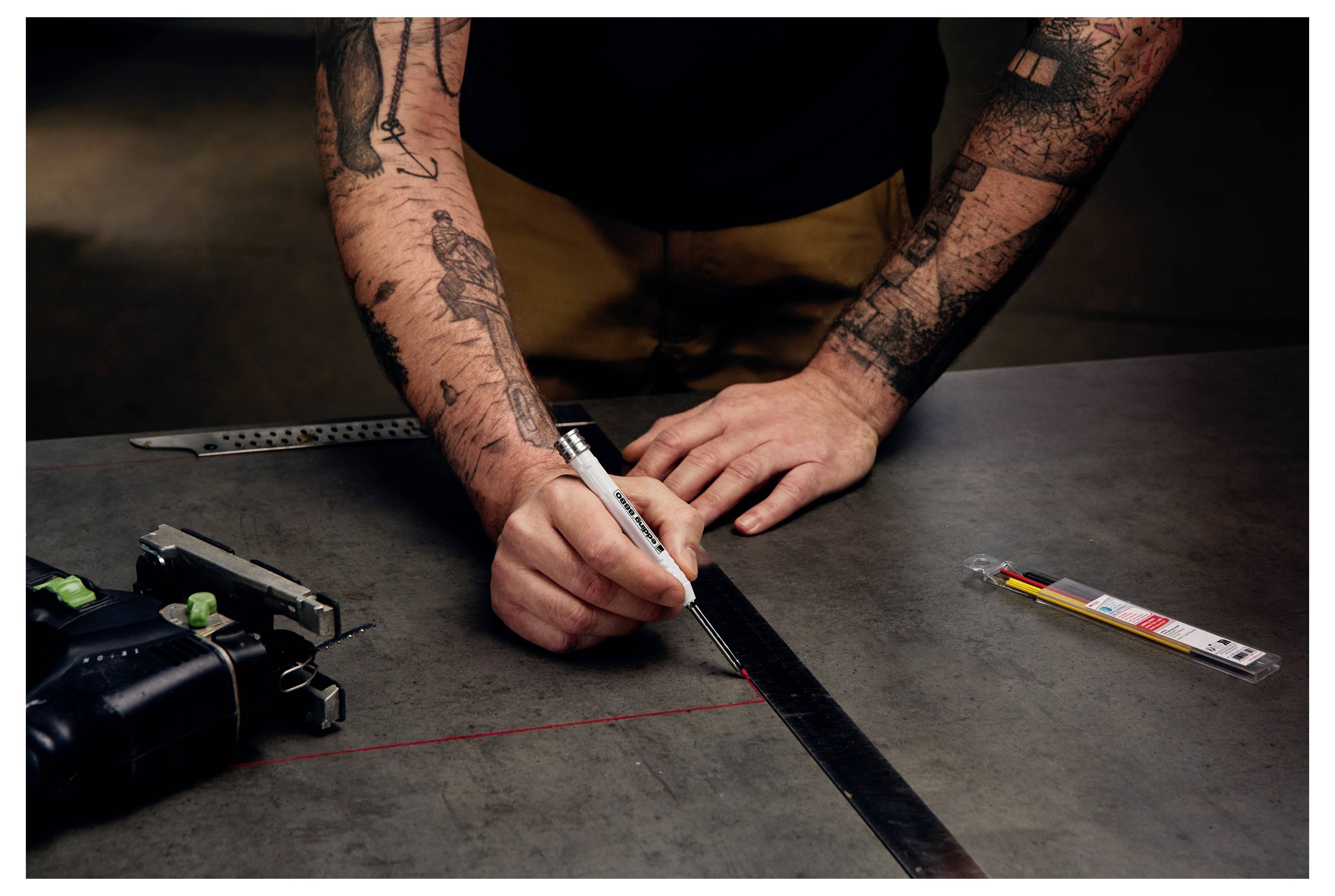Tätowierte Person, die ein Metallblech mit einem Stift und einem Stahllineal an einer Werkbank markiert, was auf Metallfertigung oder Messung hindeutet.