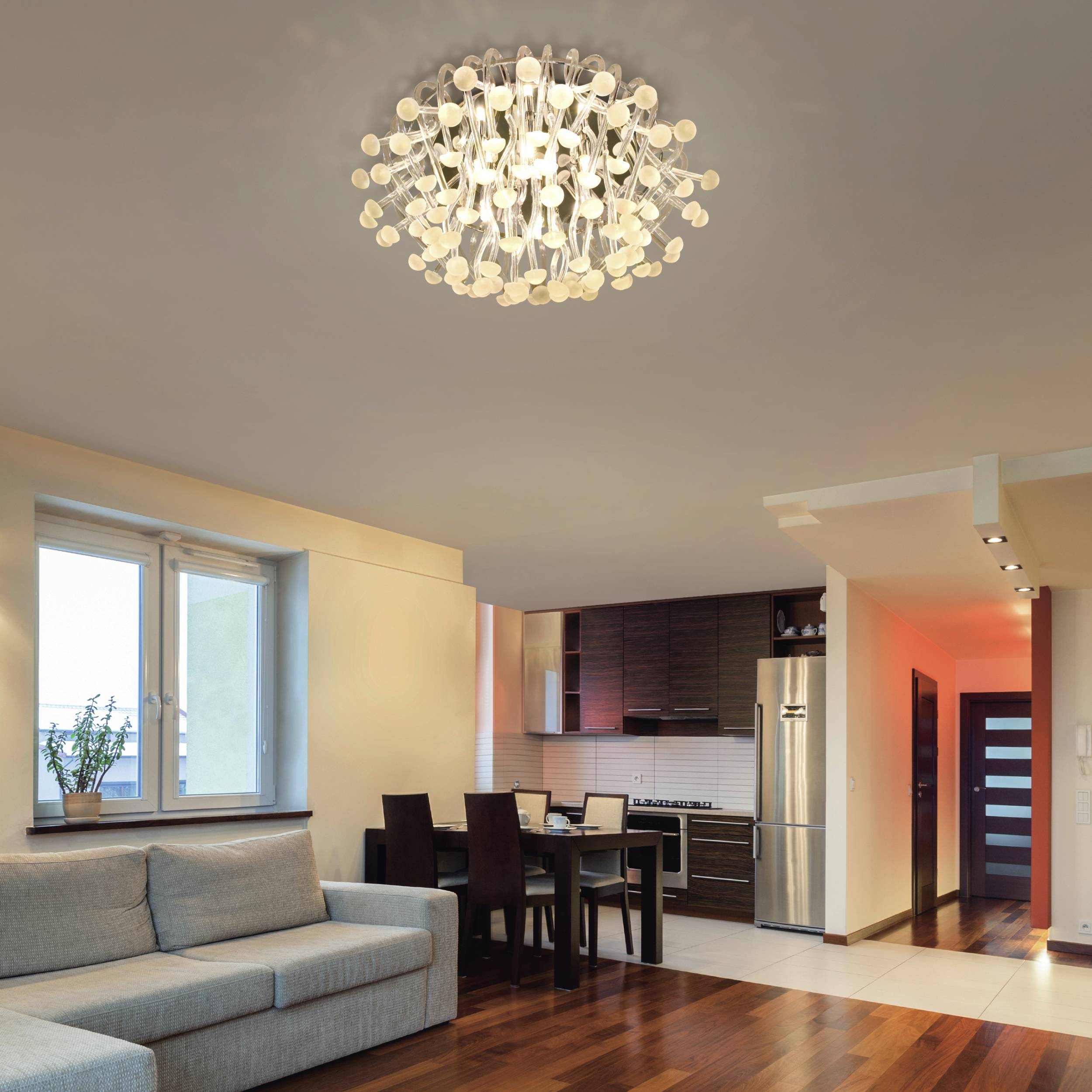 Ein modernes Wohnzimmer mit grauem Sofa, Holzboden und einem Kronleuchter. Im Hintergrund eine Küche mit dunklen Holzmöbeln.