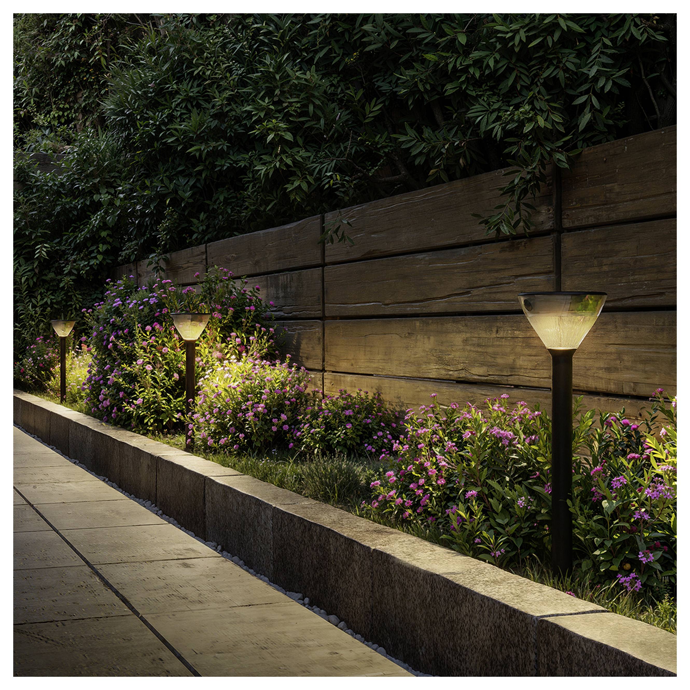 Abendlicher Gehweg mit beleuchteten Gartenweg-Leuchten, rosa Blumen und einer Holzstützmauer neben einem gepflasterten Weg.