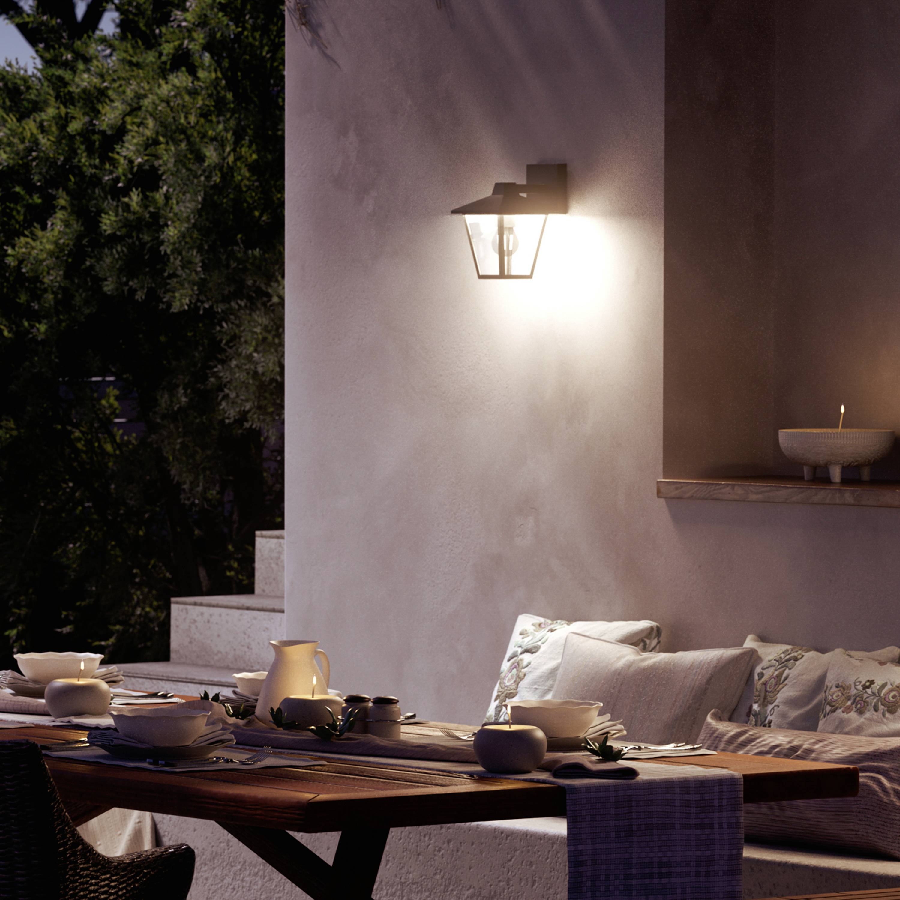 Gemütliche Terrasse bei Abenddämmerung, Tisch gedeckt mit Geschirr, Beleuchtung durch Wandlampe; ruhige, einladende Atmosphäre.