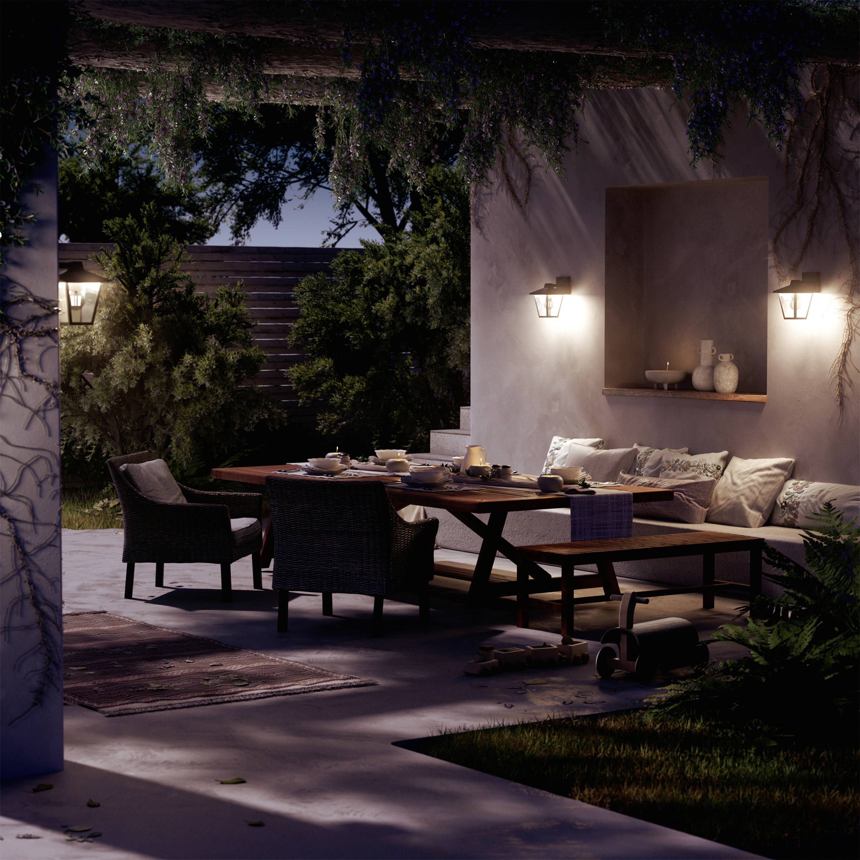 Eine gemütliche Terrasse bei Nacht, beleuchtet von Wandlampen. Ein Esstisch mit Geschirr und vier Stühlen steht im Mittelpunkt, umgeben von Pflanzen und einem Sofa.