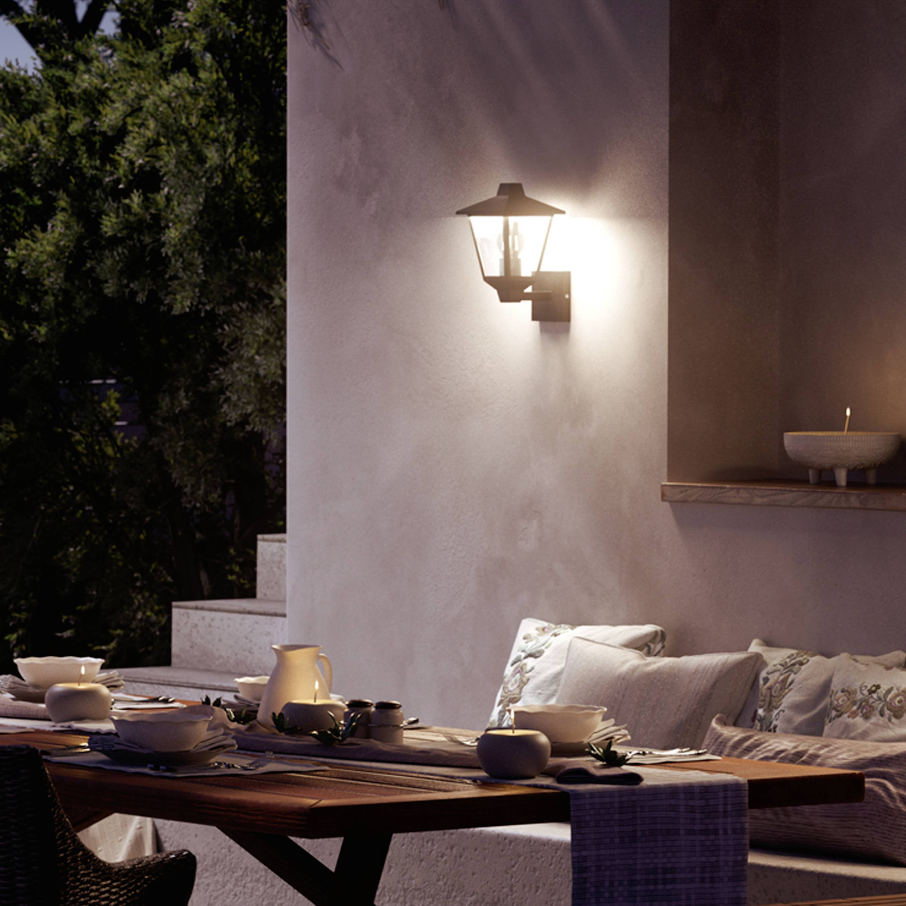 Ein Wohnzimmer im Freien mit Tisch, gedeckt für ein Abendessen. Eine Lampe an der Wand strahlt warmes Licht aus, umgeben von Bäumen.