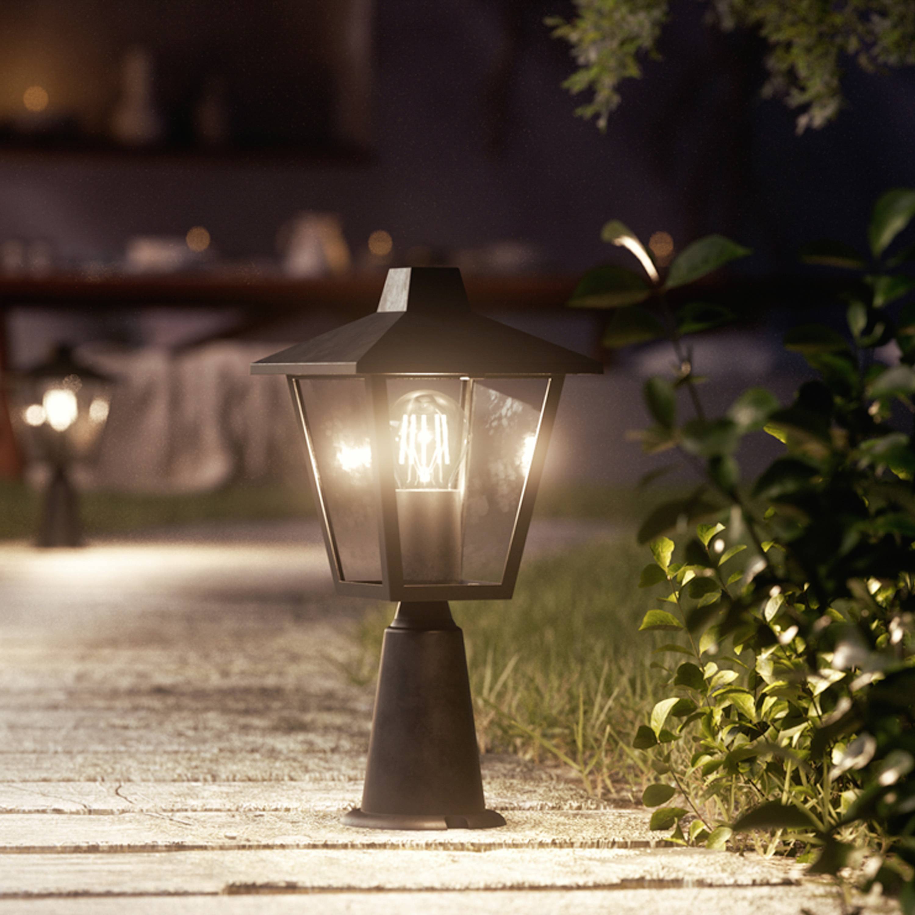 Eine beleuchtete Laterne steht auf einem gepflasterten Weg im Garten bei Nacht, umgeben von Pflanzen. Im Hintergrund ist ein gedeckter Tisch zu sehen.
