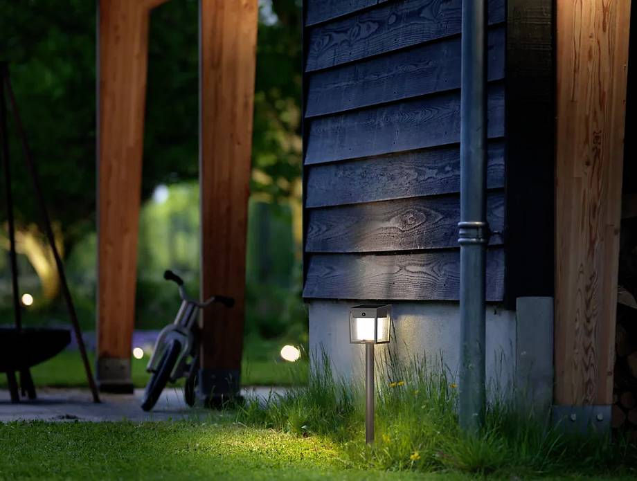 Garten bei Dämmerung mit beleuchteter Lampe, hölzernem Haus, Fahrrad und grünem Rasen. Stimmungsvolle Abendatmosphäre.