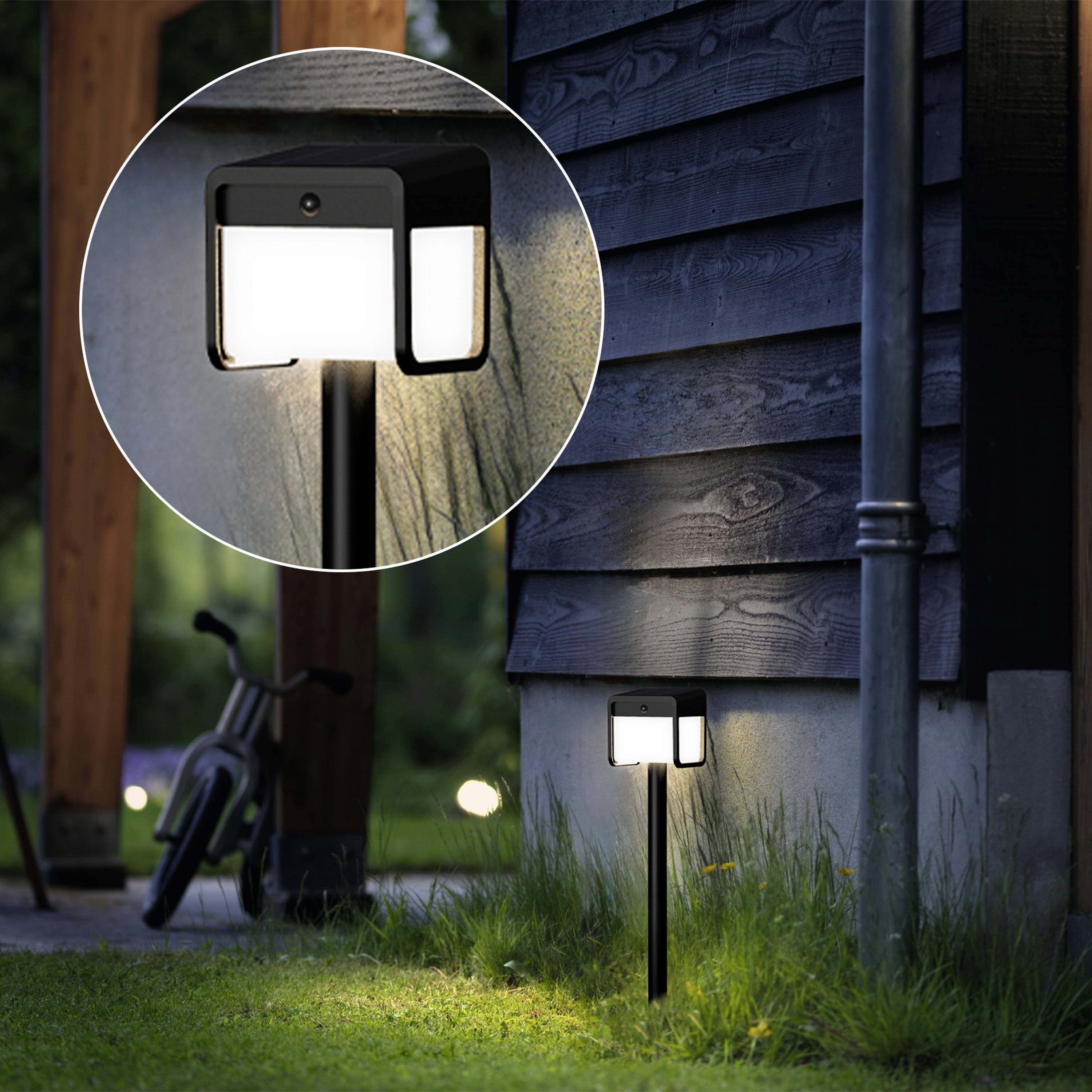 Gartenlandschaft mit einer leuchtenden Solarlampe im Vordergrund, montiert an einem Holzpfosten, neben einem Haus in der Dämmerung.