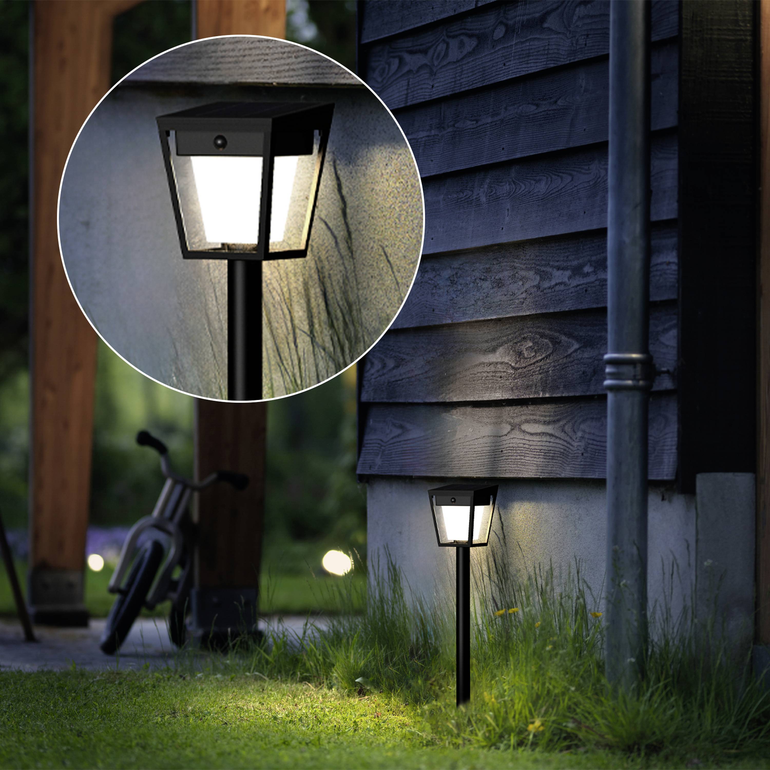 Eine leuchtende Außenlampe neben einer Holzwand, beleuchtet den umgebenden Gartenbereich. Im Hintergrund ist ein Fahrrad sichtbar.
